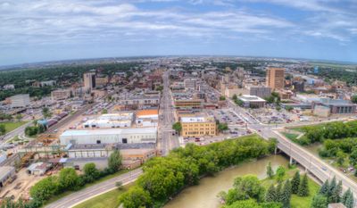 Aerial view of a city with a river curving through a lush, green park area. Buildings and streets stretch into the distance under a partly cloudy sky. A bridge crosses the river, connecting different parts of the city.