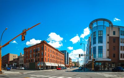 Street view with a red brick building on the left and a modern glass building on the right under a clear blue sky. Traffic lights and a few parked cars are visible on the road.