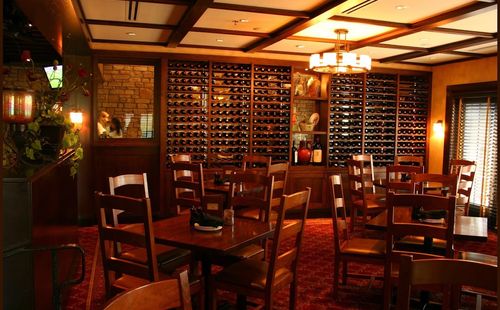Cozy restaurant interior with wooden tables and chairs, a wall lined with wine racks, dim lighting, and decorative vases. A chandelier hangs from the ceiling, and a window with closed blinds is on the right.