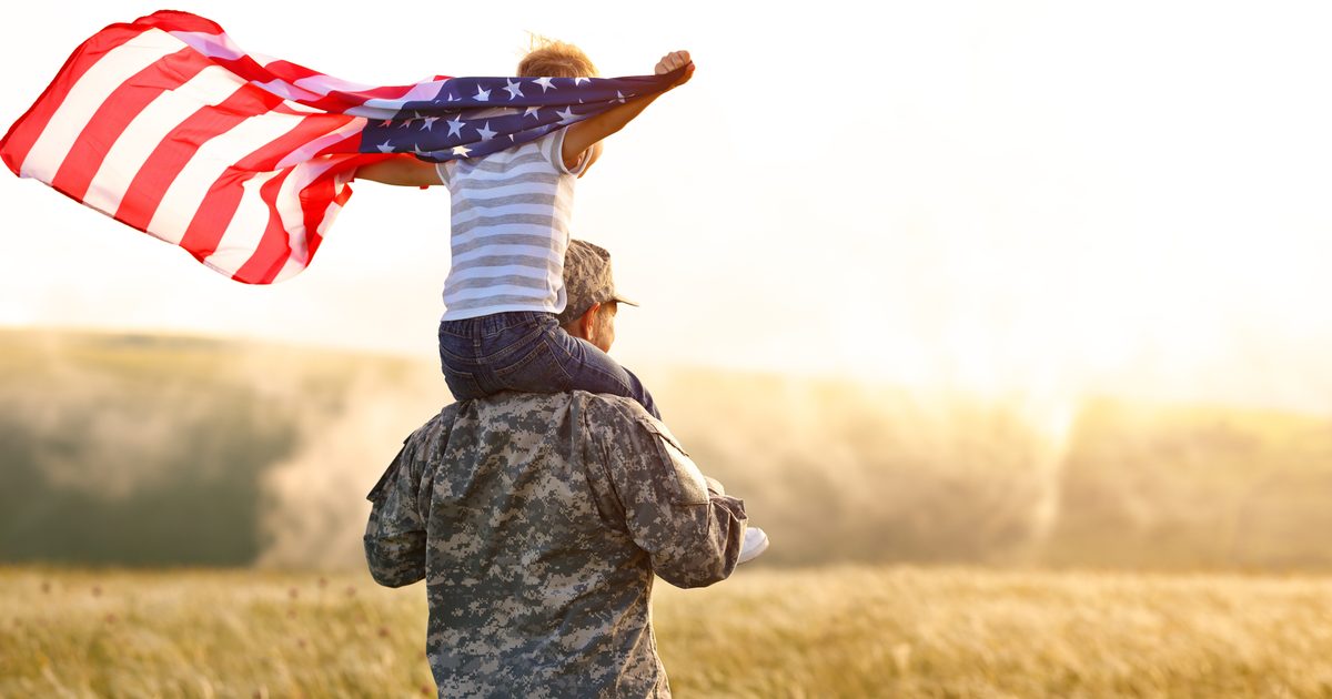 A man in a military uniform carries a child on top of his shoulders who is holding on to an American flag.