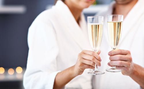 Two people in white robes clinking champagne glasses. The focus is on their hands and the glasses, with the background softly blurred.
