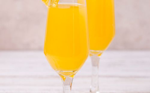 Two champagne flutes filled with orange juice, each garnished with an orange slice. They are placed on a light-colored cloth napkin on a white wooden surface. Additional orange wedges are beside the glasses.