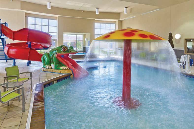 Indoor pool with a red waterslide shaped like a frog, a mushroom-shaped water fountain, and green poolside chairs. Large windows in the background provide natural light. Tile flooring surrounds the pool area.
