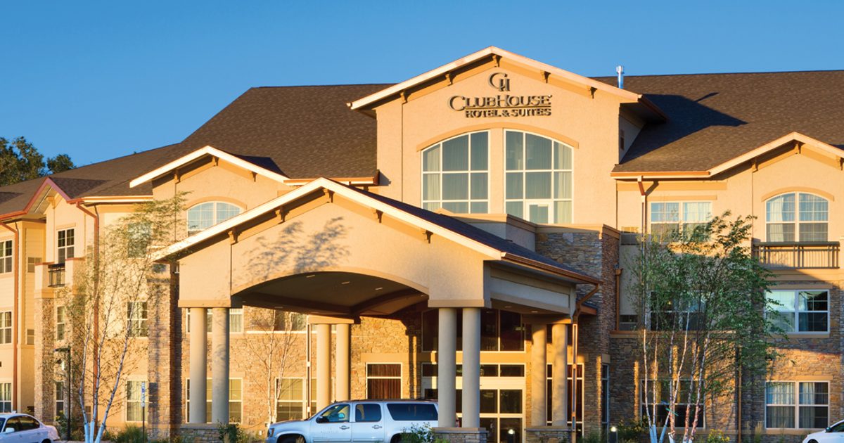 A three-story hotel building with beige and stone exterior. A covered entrance extends from the main entrance, with a white SUV parked nearby. The hotel name is visible on the front. The scene is set under a clear blue sky.