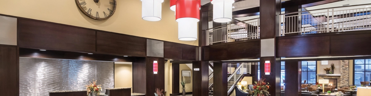 Spacious hotel lobby with modern decor. Tall ceiling with large red and white pendant lights. Stylish chairs in a circular arrangement, a textured wall behind the reception, and a large clock. Fresh flowers on tables and warm lighting throughout.
