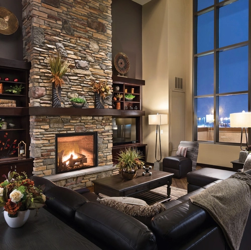 A cozy living room with a stone fireplace, black leather couches, and tall windows showing a cityscape at night. The room is decorated with plants, vases, and warm lighting. Shelves next to the fireplace display various decorative items.