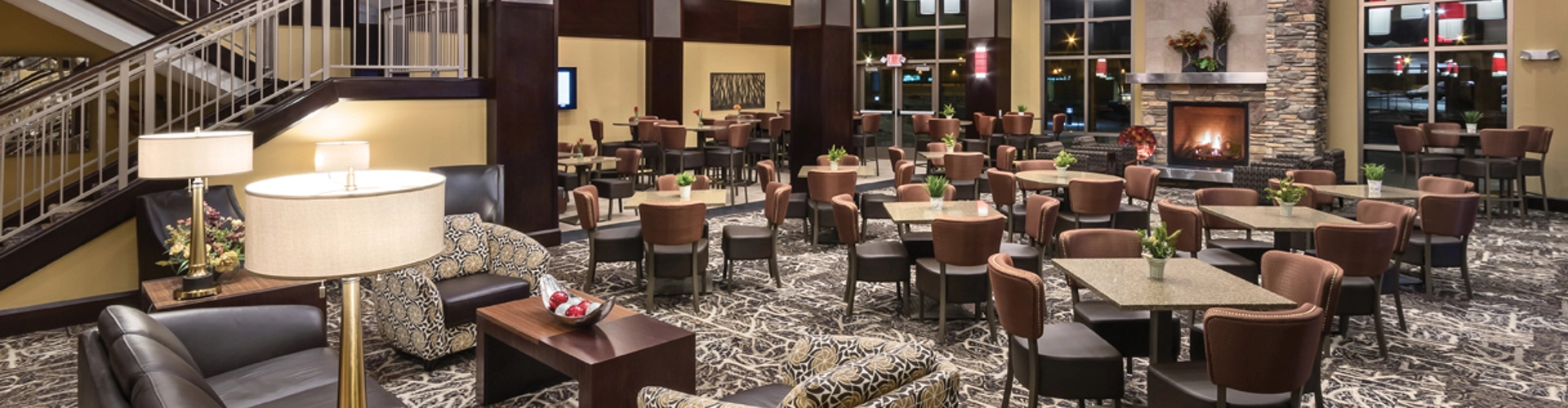 A modern hotel lobby features patterned carpet, a seating area with sofas and armchairs, and tables with chairs. Large red and white cylindrical lights hang from the ceiling. A fireplace with stone surround is at the back, near a staircase.