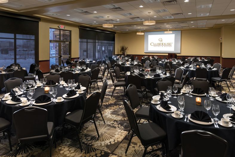 A banquet hall set up for an event. Round tables are covered with black tablecloths, set with glassware, silverware, plates, and black napkins. A projector screen at the front displays "ClubHouse Hotel & Suites." Candles and dim lighting create a warm ambiance.
