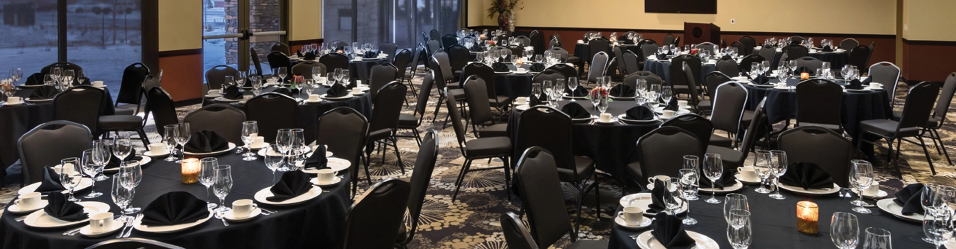 A banquet hall set for an event. Round tables covered with black tablecloths are arranged neatly, each set with glasses, plates, and black napkins. Chairs surround the tables, and dim lighting creates a formal atmosphere.