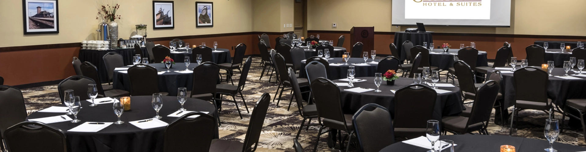 A conference room set up for an event with round tables covered in black tablecloths. Each table has place settings and notepads. A presentation screen at the front displays the "ClubHouse Hotel & Suites" logo. Dim overhead lighting creates a warm ambiance.