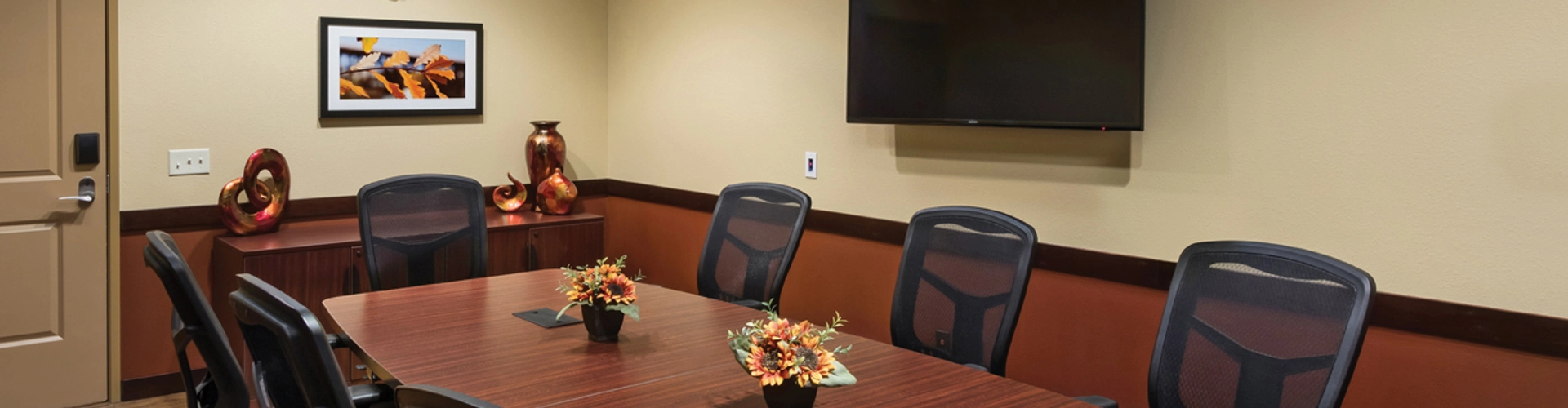 A conference room with a long wooden table surrounded by black mesh chairs. Three small floral arrangements are on the table. A wall-mounted TV and a framed picture are on the walls, and the floor is wooden.