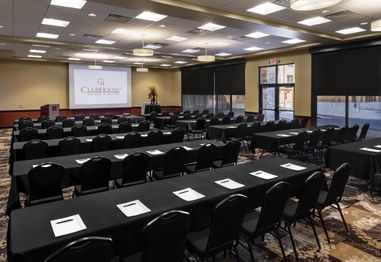 A conference room with rows of black-clothed tables and chairs facing a projector screen. Notepads and pens are placed on the tables. The room is well-lit, with large windows and ceiling lights. Walls are beige, and the carpet has a floral pattern.