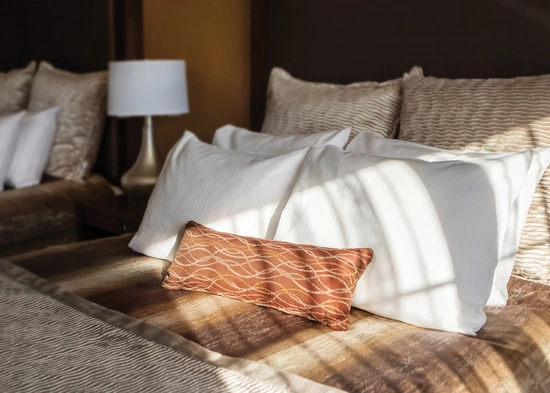A bed with two large tan pillows, four small white pillows, and one red accent pillow. The sun is coming through the window, casting shadows on the bedding. A lamp sits on a table next to the bed.