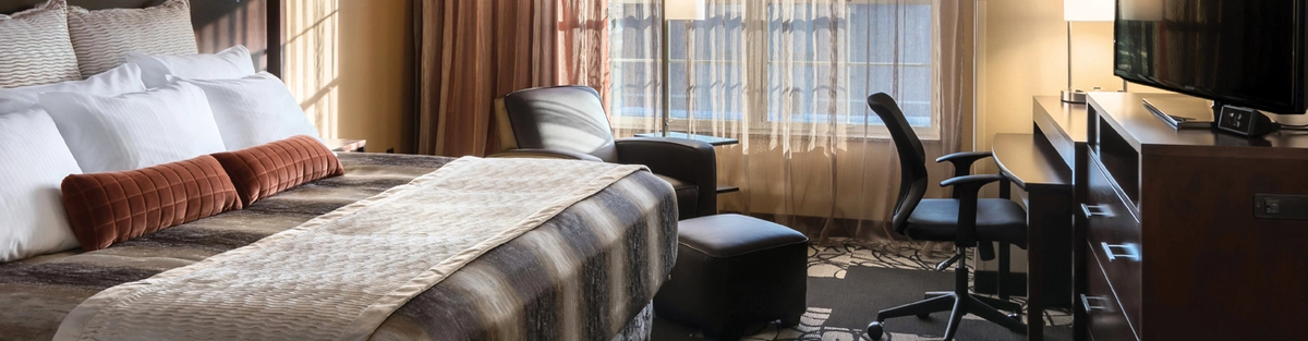 A guest room with a king bed on the left. The bedding is striped and there are two red accent pillows. On the right is a desk and TV. In the middle of the room is a chair that sits in front of the window.