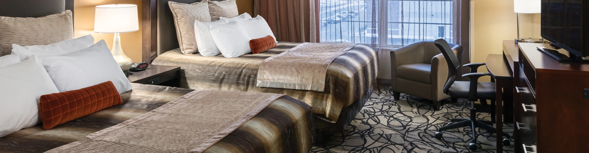 A guest room with two queen beds. Each bed has a striped brown and tan cover with a red accent pillow. The carpet is gray and tan with a black swirled pattern. The window shades are open revealing a parking lot and another building.